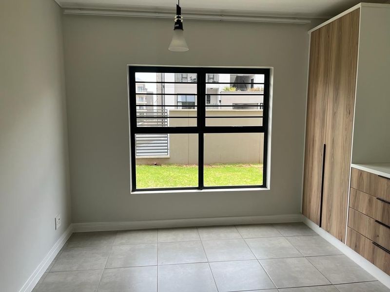 A bright, modern bedroom featuring neutral tiled flooring, built-in wood-finish cupboards, and a large window with security bars overlooking a garden area.