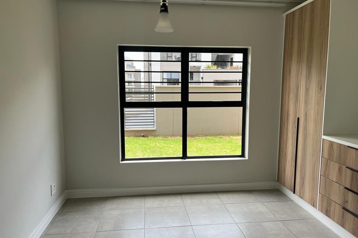 A bright, modern bedroom featuring neutral tiled flooring, built-in wood-finish cupboards, and a large window with security bars overlooking a garden area.