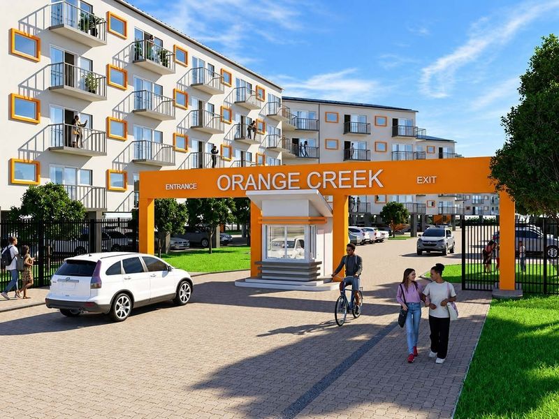 A modern residential complex entrance featuring a security gatehouse, paved driveway, and multi-story apartment buildings with balconies.