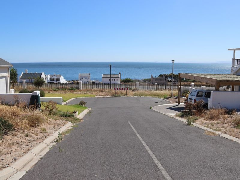 A street-level view of a residential access road leading towards a coastal area with ocean views and surrounding residential properties.