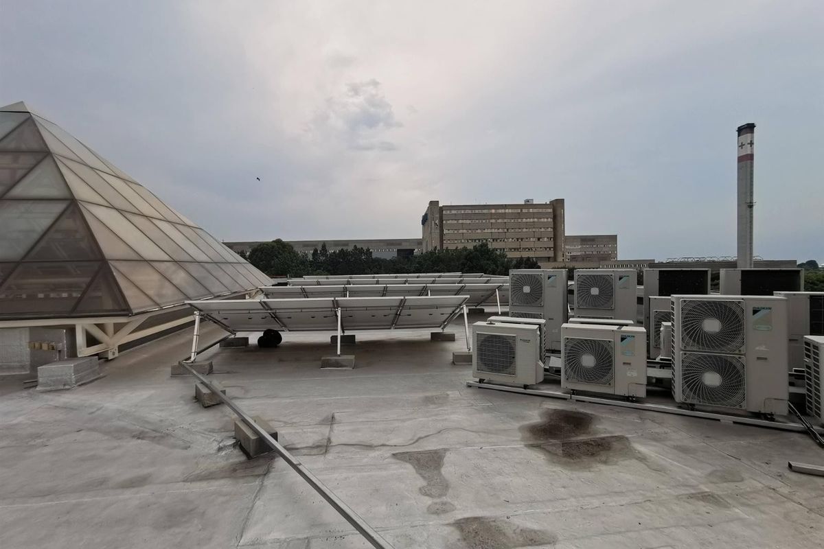 Commercial rooftop area featuring a large glass pyramid skylight, multiple HVAC condenser units, and an array of solar panels on a concrete deck.
