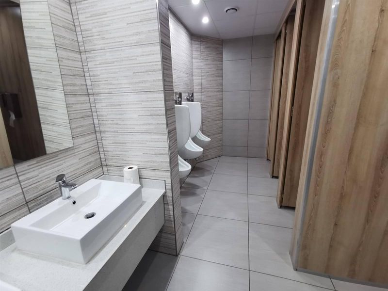 Modern commercial restroom featuring tiled walls, stone vanity with vessel sink, multiple wall-mounted urinals, and private partitioned toilet cubicles.
