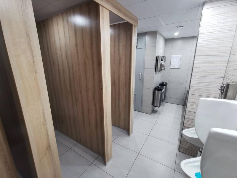 A clean, modern commercial restroom featuring tiled surfaces, multiple toilet cubicles with wood-look partitions, and wall-mounted urinals.