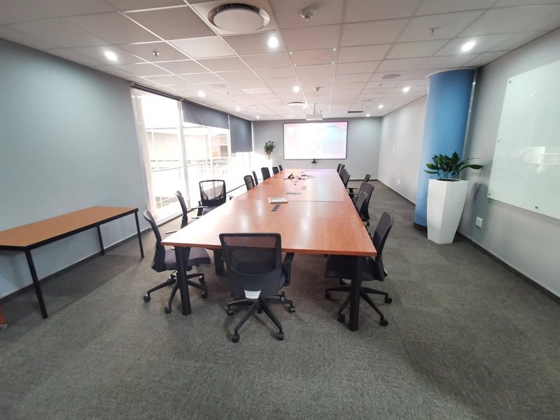 A professional commercial boardroom featuring a large conference table, suspended ceiling with recessed lighting, projector screen, and a wall-mounted glass whiteboard.