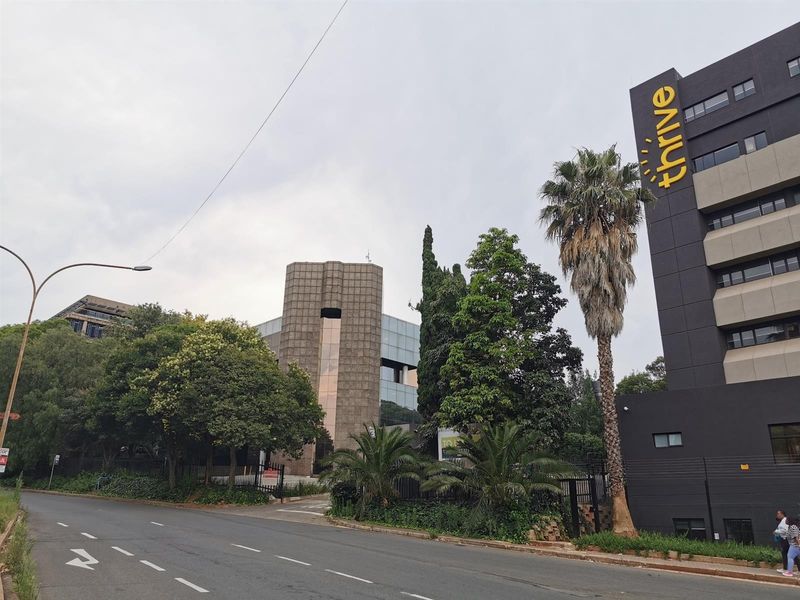 Street-level view of a commercial office building complex featuring modern facade architecture, mature landscaping, and secure perimeter fencing.