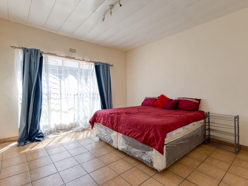 A spacious bedroom featuring neutral walls, tiled flooring, and natural light from a large window with curtain fixtures.