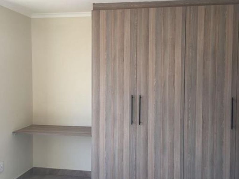 A clean bedroom featuring modern wood-grain built-in cupboards, a matching floating desk, and neutral tiled flooring.
