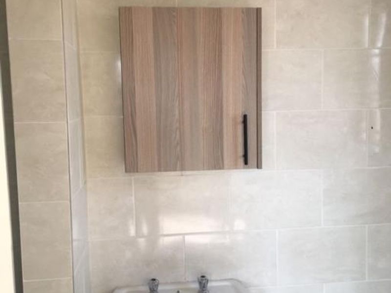 A clean bathroom featuring a white pedestal sink and a modern wood-finish wall-mounted medicine cabinet against neutral tiled walls.