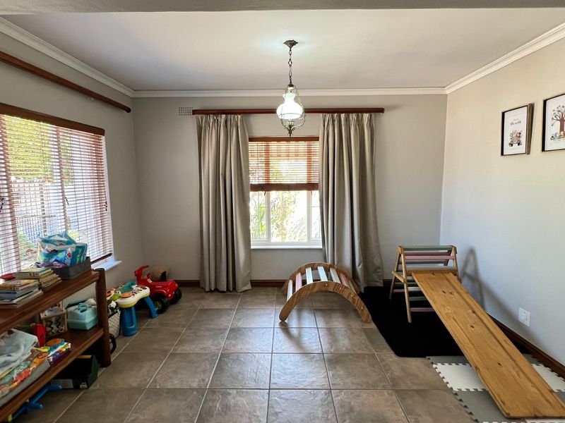 A bright living area featuring neutral walls, tiled flooring, and large windows with timber blinds, offering a versatile space for family living.