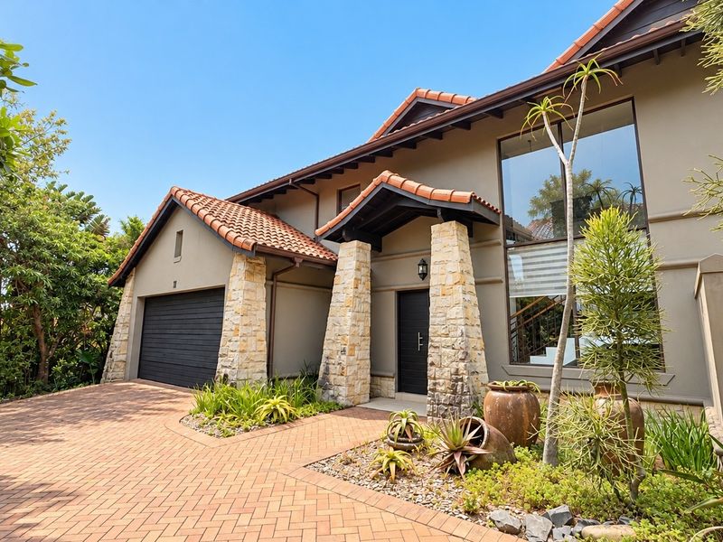 A well-maintained front facade featuring stone-clad pillars, terracotta roof tiles, a paved driveway, and a double garage.