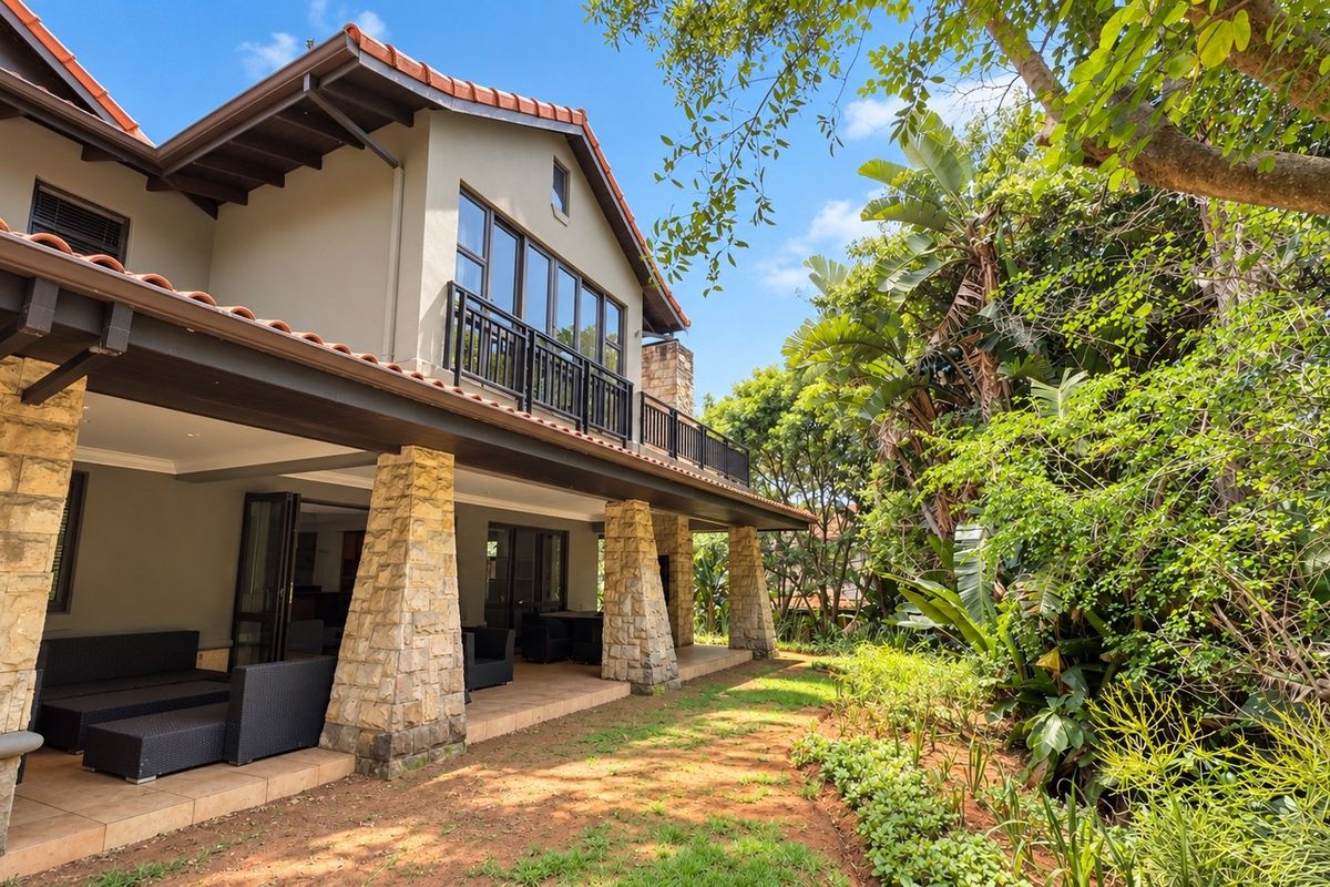 A well-maintained rear exterior featuring a spacious covered patio with stone-clad pillars, a balcony, and lush garden surroundings.