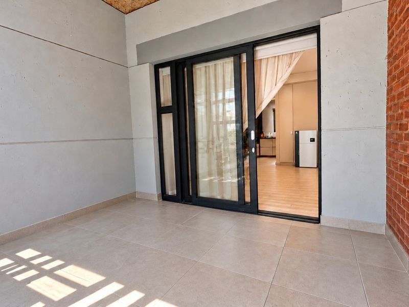 A modern, covered balcony featuring tiled flooring, an exposed brick feature wall, and large sliding glass doors providing access to the interior living space.