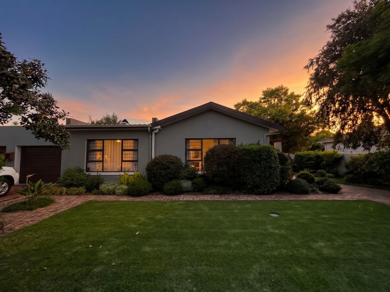 A well-maintained single-story residential home featuring a neat front garden, brick-paved driveway, and modern aluminum-framed windows at sunset.
