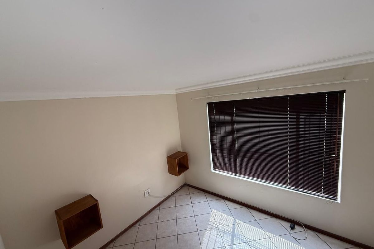 A bright, empty bedroom featuring neutral walls, tiled flooring, and wall-mounted shelving units, complemented by dark venetian blinds.