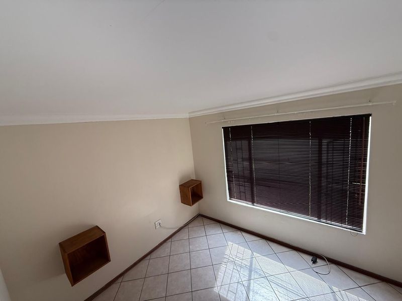 A bright, empty bedroom featuring neutral walls, tiled flooring, and wall-mounted shelving units, complemented by dark venetian blinds.