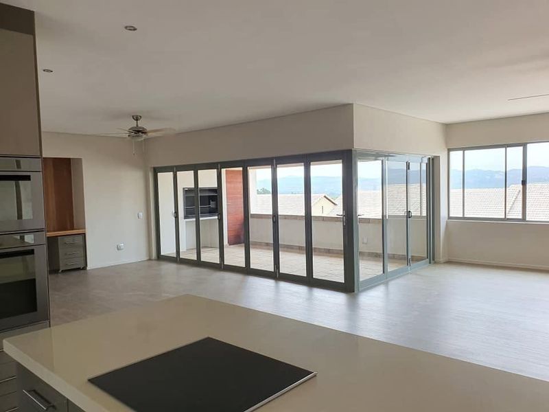 Spacious open-plan living area featuring a modern kitchen with built-in appliances, flowing seamlessly through stacker doors to a covered patio with a built-in braai.