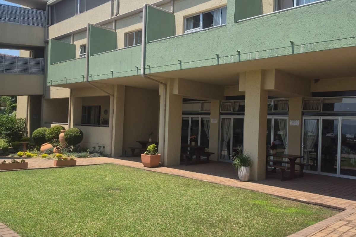 Rear view of a retirement village building featuring a paved patio area, ground floor access via sliding doors, and upper-level balconies overlooking a landscaped garden.