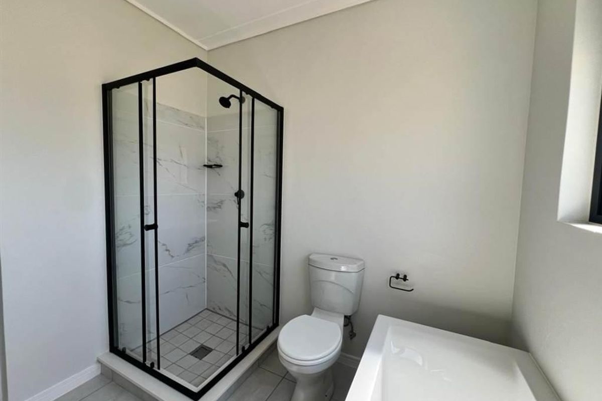 A modern, clean bathroom featuring a glass-enclosed shower with black framing, a white toilet, and a bathtub with neutral tiled flooring.
