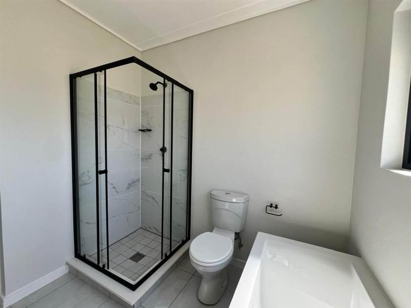 A modern, clean bathroom featuring a glass-enclosed shower with black framing, a white toilet, and a bathtub with neutral tiled flooring.