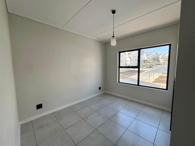 A bright, empty bedroom featuring neutral grey floor tiles, a modern black-framed window, and a simple pendant light fixture.