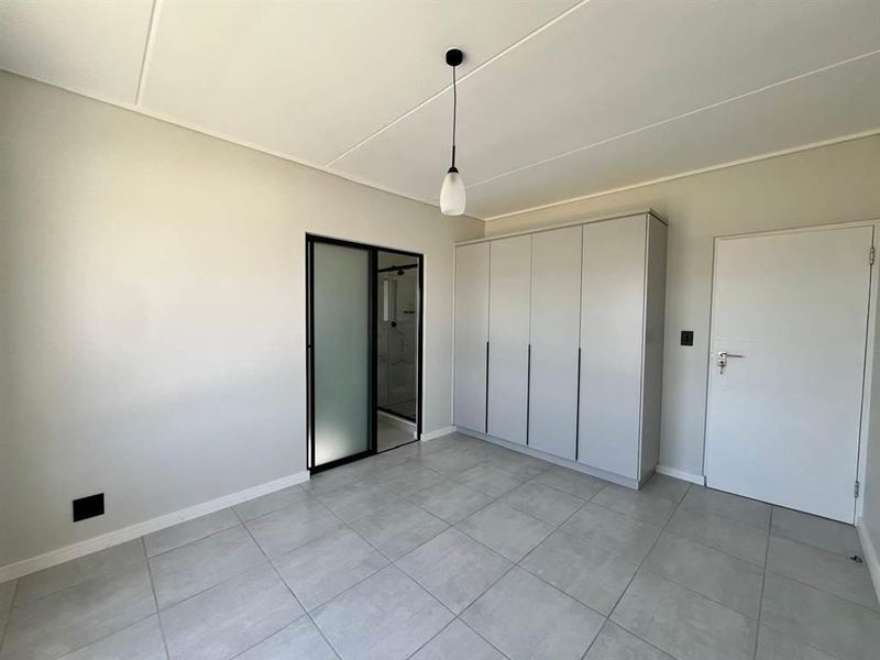 A clean, modern bedroom featuring neutral tiled flooring, a large built-in wardrobe, and a sleek frosted glass door leading to an en-suite bathroom.