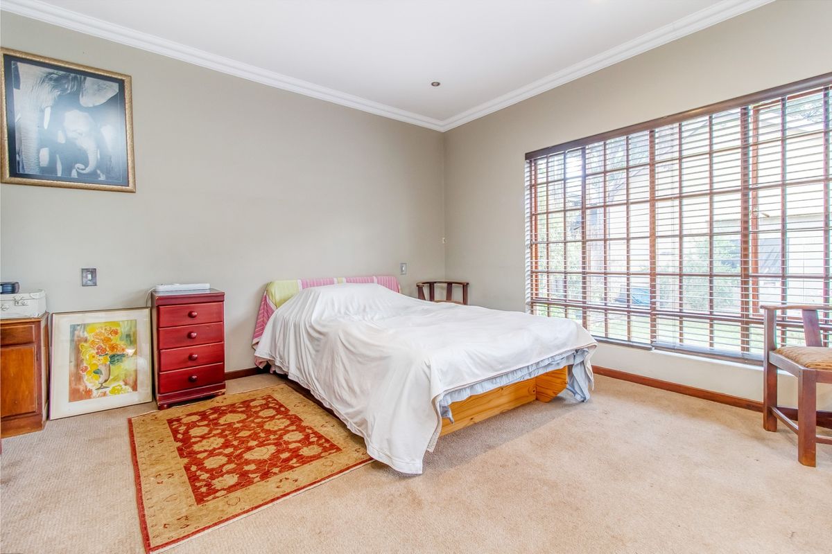A well-lit bedroom featuring neutral wall tones, carpeted flooring, and a large window fitted with venetian blinds.