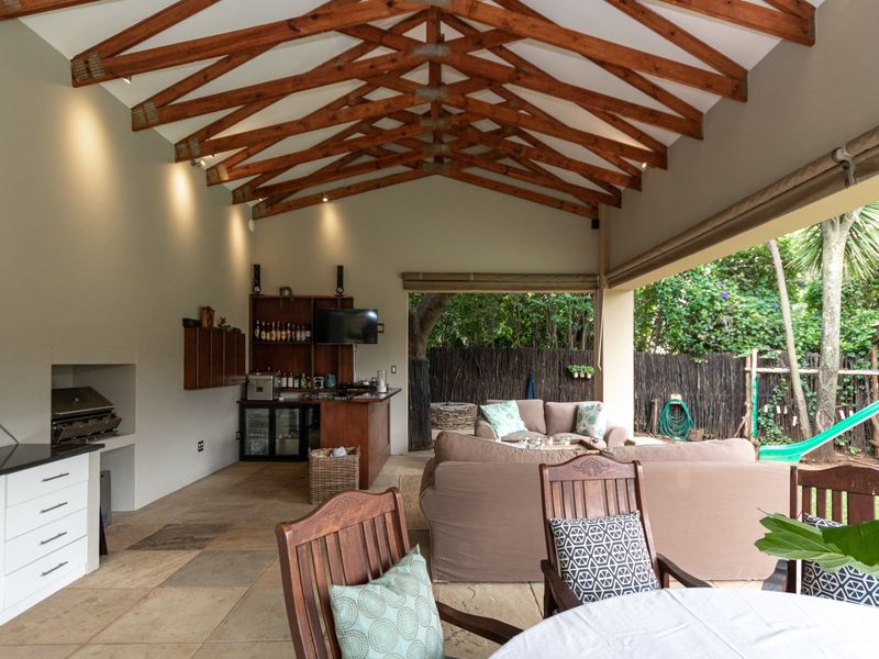 Spacious covered patio featuring exposed timber roof trusses, stone tile flooring, built-in braai, and integrated cabinetry, seamlessly connecting to the garden area.