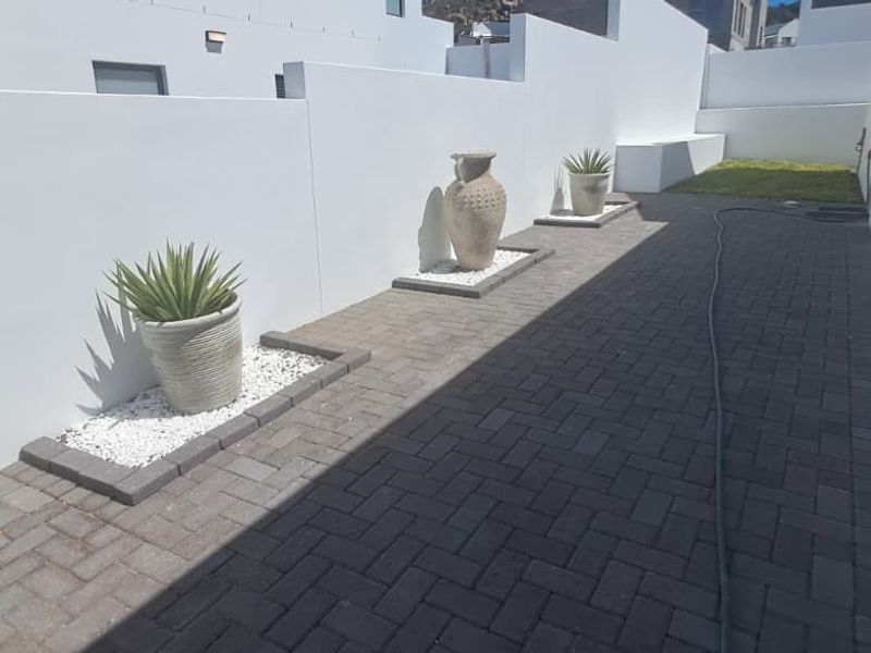 A clean, modern rear exterior area featuring a paved walkway, white boundary walls, and landscaped garden beds with a small lawn section.
