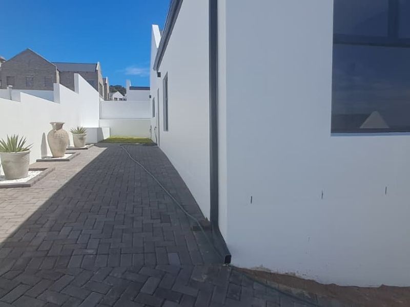 A side exterior view of the property featuring a paved walkway, white boundary walls, and modern aluminium window frames.