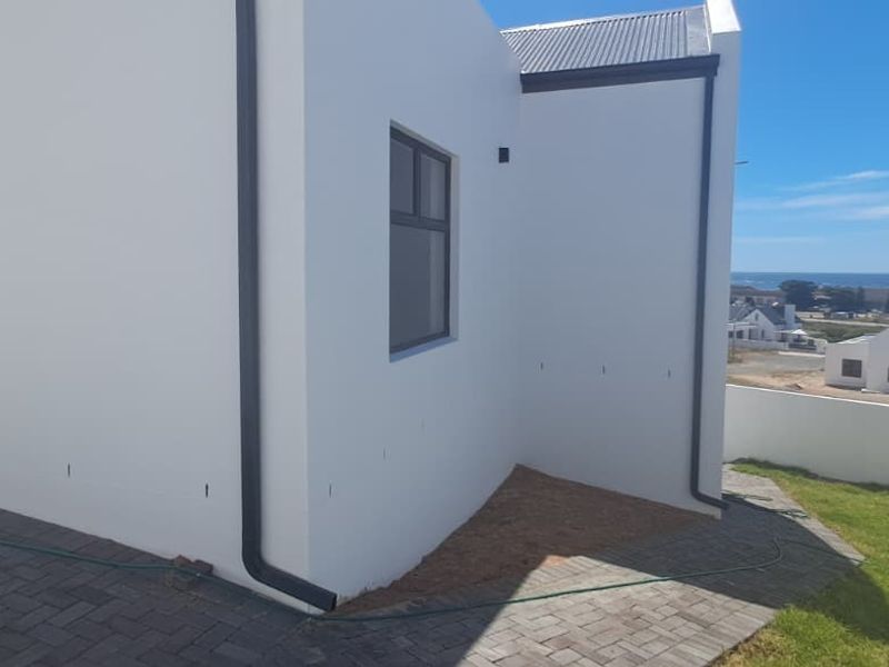 Rear exterior view of the property featuring a paved area, white rendered walls, modern downpipes, and a view towards the coastline.