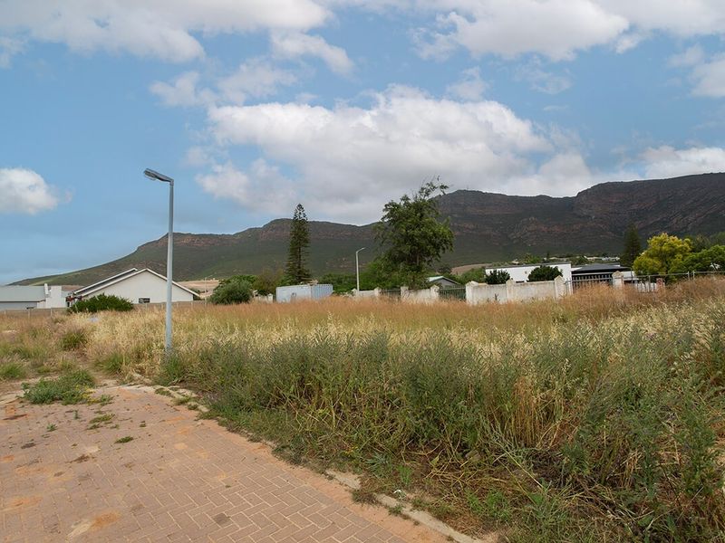 A vacant residential plot featuring a paved driveway, perimeter fencing, and street lighting, set against a scenic mountain backdrop.