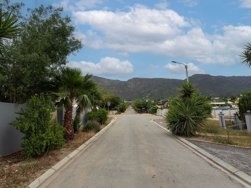 A clear street view showing the paved road, concrete curbing, and surrounding residential boundary walls with mountain views in the background.