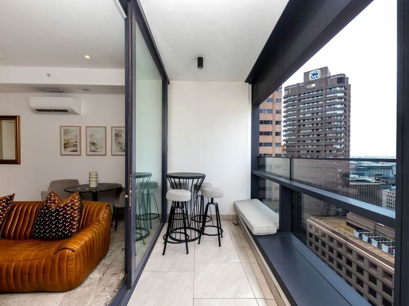 A modern, covered balcony featuring tiled flooring, a glass balustrade, and built-in bench seating, offering an elevated urban view.