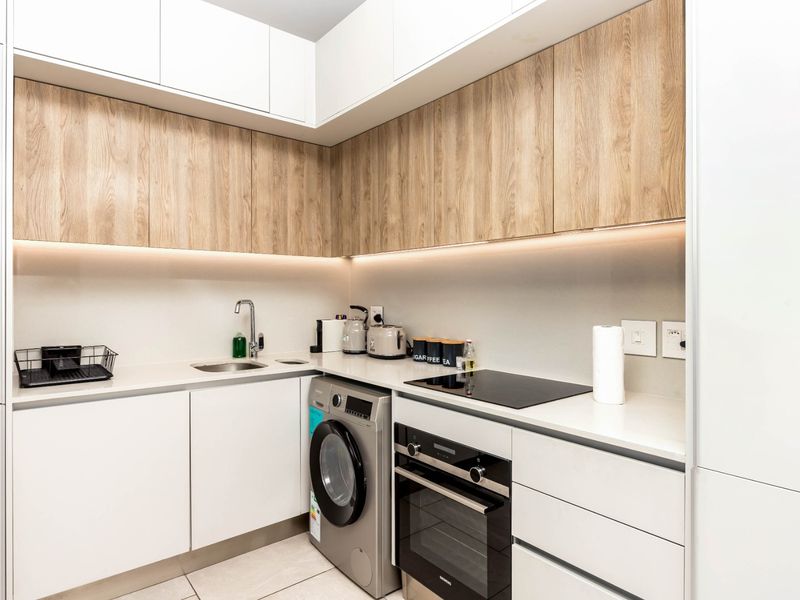 Modern compact kitchen featuring sleek white and wood-grain cabinetry, stone countertops, integrated appliances, and efficient under-cabinet lighting.