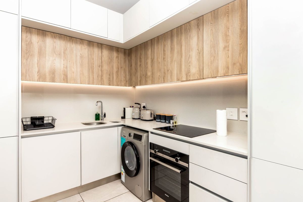 Modern compact kitchen featuring sleek white and wood-grain cabinetry, stone countertops, integrated appliances, and efficient under-cabinet lighting.