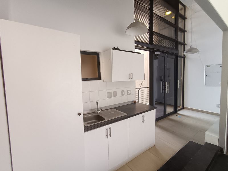 A compact kitchenette featuring white cabinetry, a stainless steel sink, tiled backsplash, and modern pendant lighting, situated near a large glass entrance door.