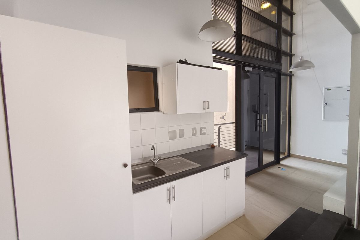A compact kitchenette featuring white cabinetry, a stainless steel sink, tiled backsplash, and modern pendant lighting, situated near a large glass entrance door.