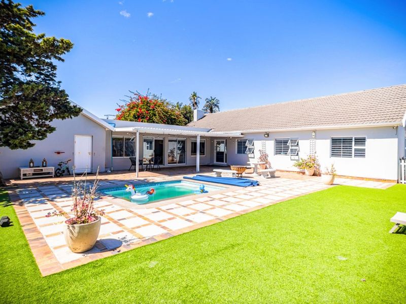 Rear exterior view featuring a swimming pool, paved pool deck, covered patio area, and well-maintained artificial lawn.