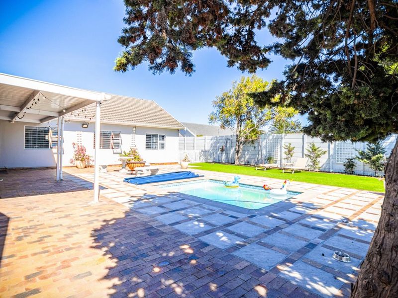A well-maintained backyard featuring a swimming pool, covered patio area, brick paving, and artificial lawn, enclosed by a perimeter wall.