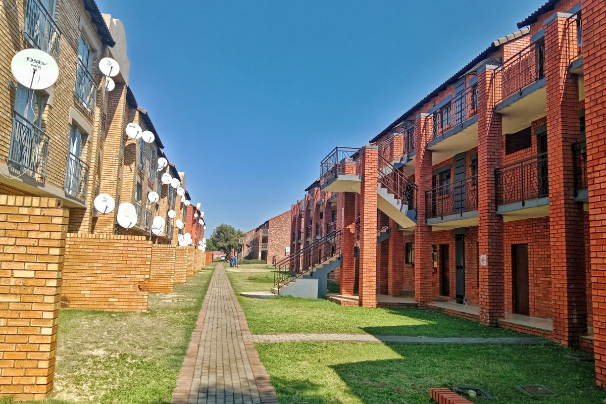 Exterior view of a multi-story apartment complex featuring face brick construction, external staircases, balconies, and a central paved walkway through a communal garden area.