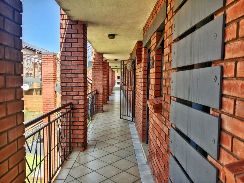 An exterior apartment hallway featuring exposed brick columns, tiled flooring, and metal security railings, providing access to individual units.