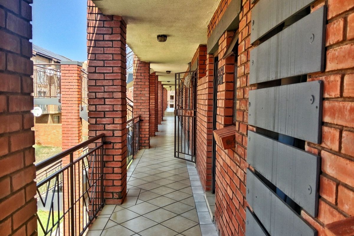An exterior apartment hallway featuring exposed brick columns, tiled flooring, and metal security railings, providing access to individual units.