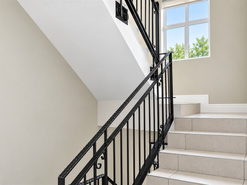 A bright and modern staircase featuring tiled steps and a decorative black wrought iron balustrade, illuminated by a large window.