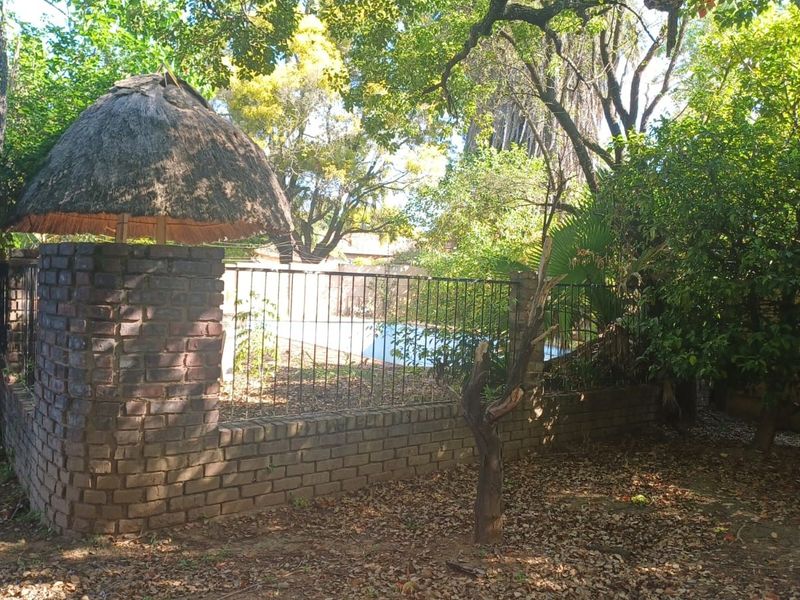 Outdoor pool area featuring a brick boundary wall, metal fencing, and a traditional thatched lapa, surrounded by mature trees and garden foliage.