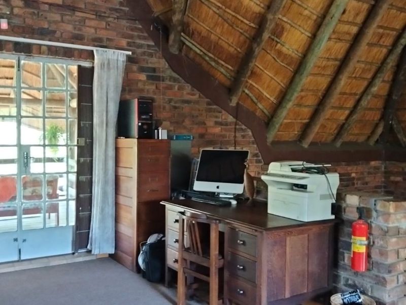 A rustic home office featuring exposed brick walls, a traditional thatched roof with timber rafters, and a glass-paneled door leading to an outdoor area.