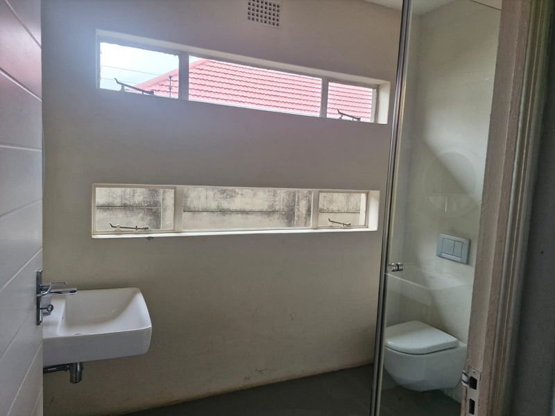 A minimalist bathroom featuring a wall-mounted basin, wall-hung toilet with concealed cistern, and a glass shower enclosure, illuminated by horizontal casement windows.