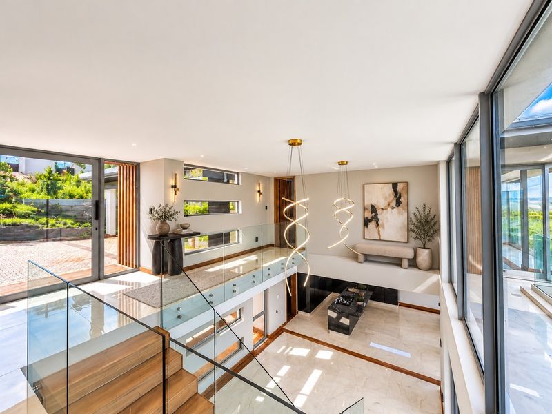 A modern, double-volume living area featuring a glass balustrade, timber staircase, and expansive floor-to-ceiling windows that provide abundant natural light and garden views.