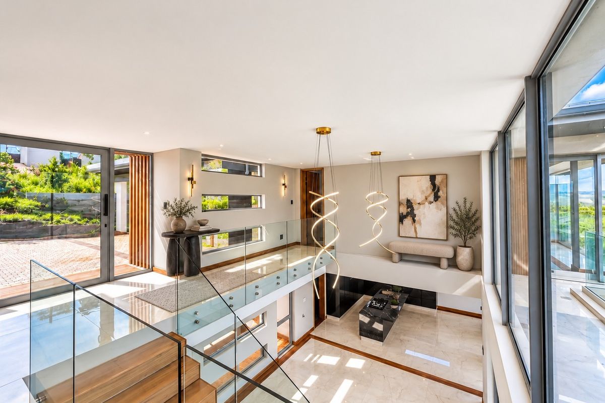 A modern, double-volume living area featuring a glass balustrade, timber staircase, and expansive floor-to-ceiling windows that provide abundant natural light and garden views.