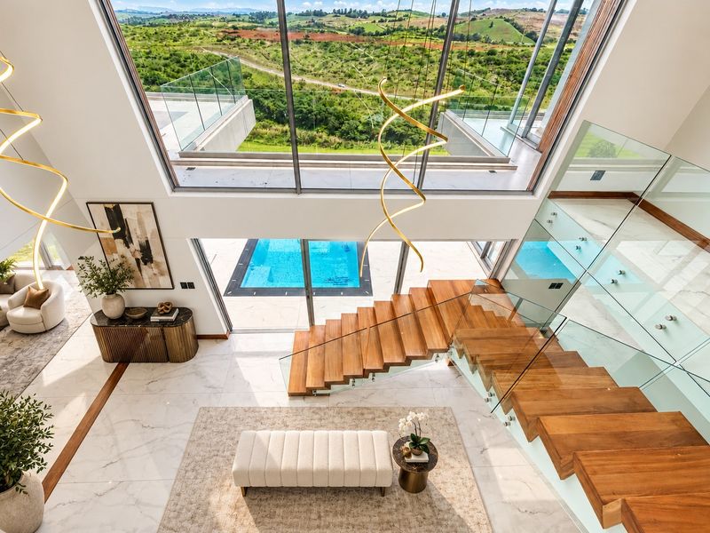 A luxurious double-volume living area featuring a floating timber staircase, marble flooring, and expansive floor-to-ceiling windows overlooking a pool and scenic landscape.
