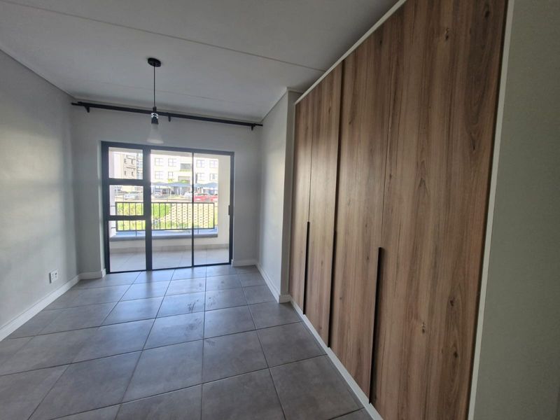 A bright, empty bedroom featuring modern wood-finish built-in cupboards, tiled flooring, and a sliding glass door leading to a balcony.